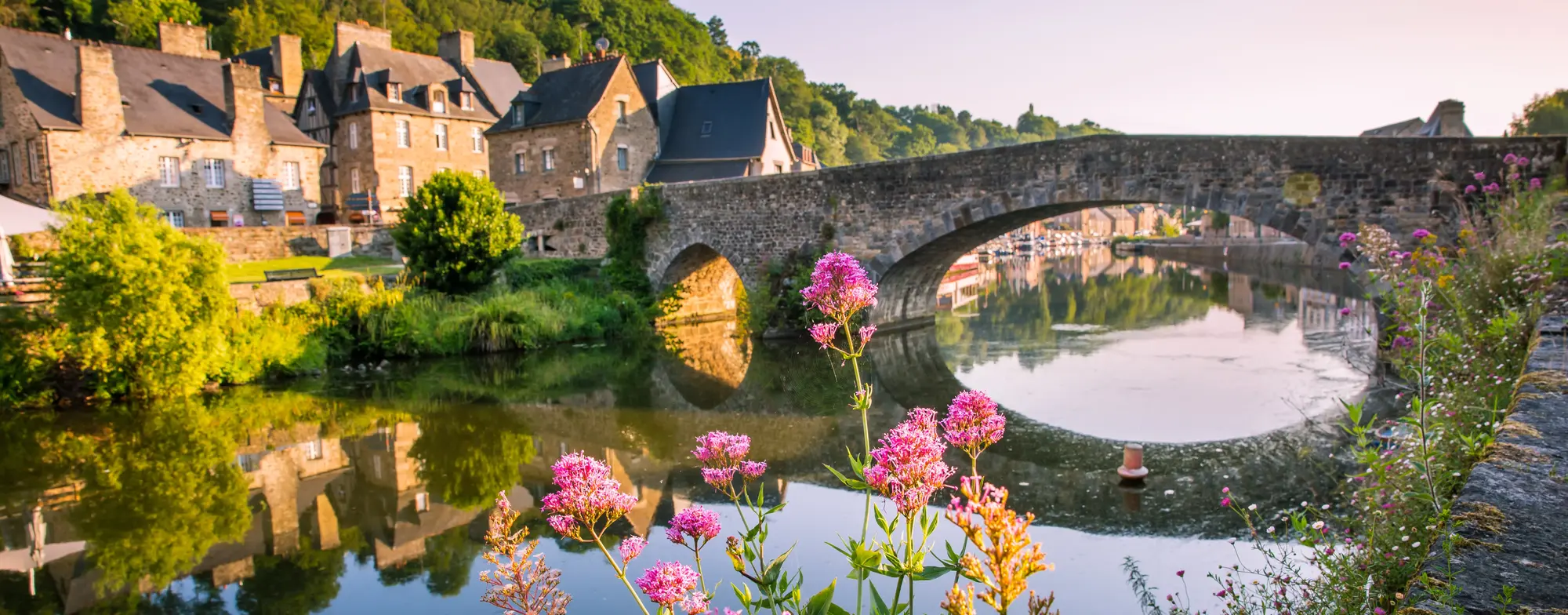 Cotes d'Amor, Bretagne - Ein Foto von einer Brücke über einem Fluss, im Vordergrund Pinke Blumen und links vom Bild, am Rand des Flusses befinden sich Büsche, welche sich im Wasser spiegeln. Im Hinergrund sieht man Häuser und einen hellen Himmel.